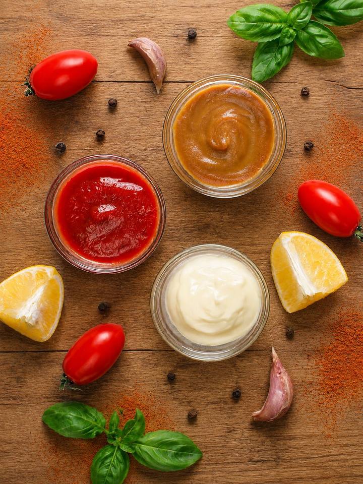 Três potinhos de molho para salada sobre mesa de madeira, com tomate, limão, alho e manjericão