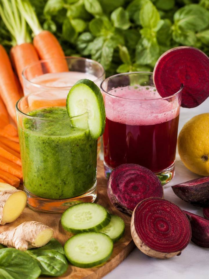 Copos de suco verde e de suco de beterraba, com cenoura, pepino e gengibre ao lado